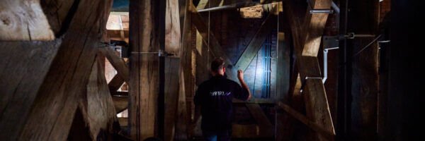 Inspecteur van Nusman met zaklamp voert een elektrotechnische inspectie uit in een houten kapconstructie.