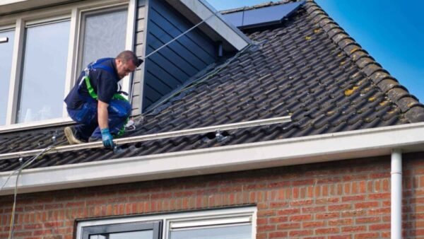Een persoon met veiligheidsuitrusting installeert zonnepanelen op het schuine dak van een huis met bakstenen gevel en grote ramen