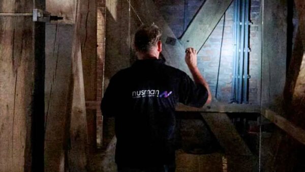 Een man in een zwart shirt staat in een kamer met balken en op de achtergrond leidingen.