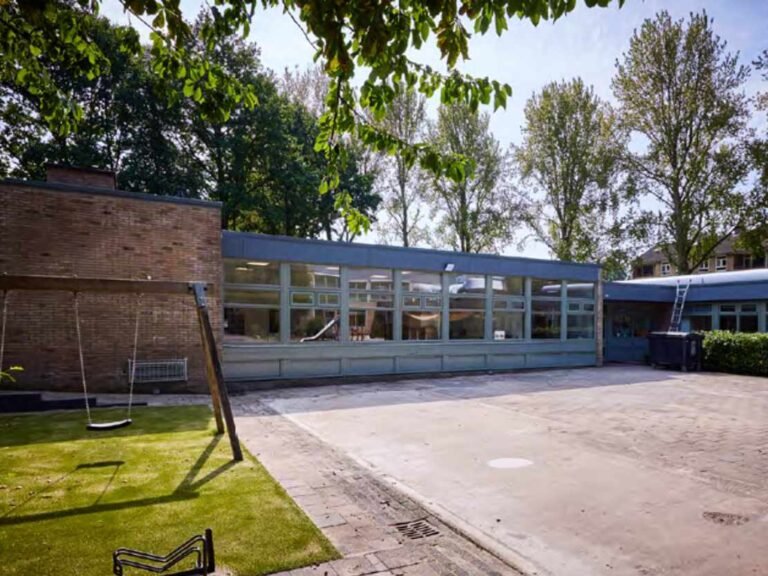 Buitenzijde van een schoolgebouw in Groningen met een betegeld schoolplein, een speelschommel op het gras en een lange glazen gevel.