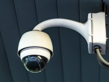 Witte beveiligingscamera met glazen koepel, gemonteerd aan een grijze muur met witte arm en kabels, voor Nusman Installatietechniek