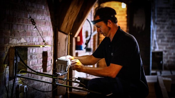 Technicus van Nusman Installatietechniek in zwart poloshirt en gele werkhandschoenen legt of onderhoudt een grijs brandbeveiligingssysteem aan in een historische bakstenen kelder met houten balken, kabels, brandblusser en warm geel licht, nabij de Martinitoren in Groningen.