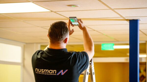 Monteur van Nusman Installatietechniek scant en controleert de werking van LED-noodverlichting in een systeemplafond.